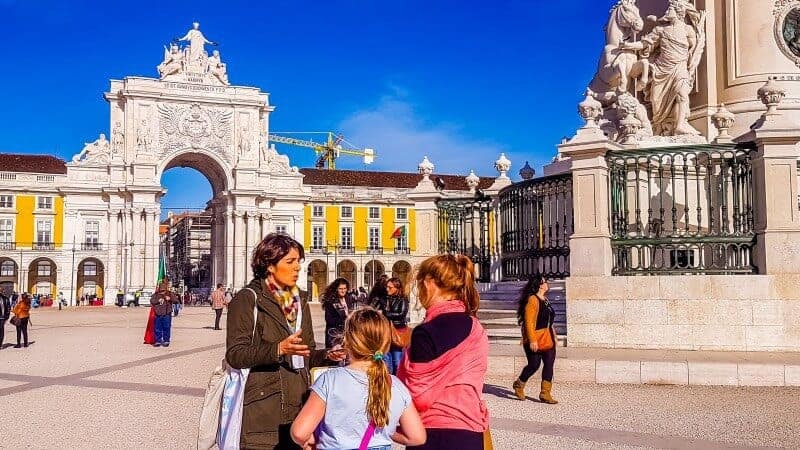 Mariana from Little Lisbon tours for kids with Willow and Marley in Commercio Square Lisbon discussing the balconies around the square and who lived in them. The yellow from the buildings is nice and bright against the blue sky