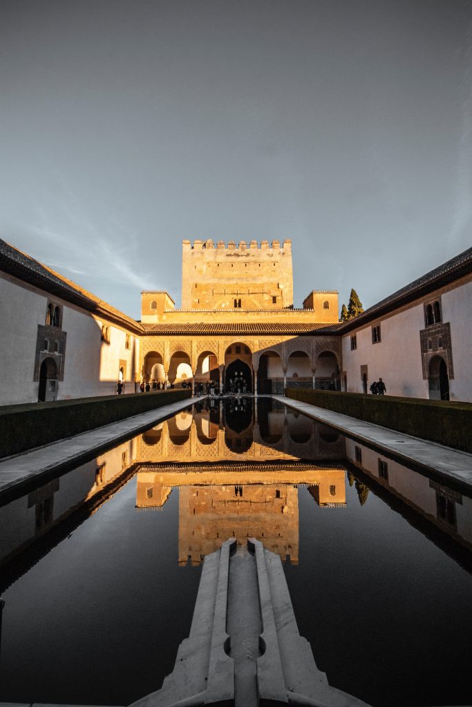One of the sandstone buildings rises over a rectangular pond in a courtyard of the Alhambra in Granada. The sun is bouncing off the sandy colours building and it is reflected perfectly in the mirror-like water of the pond