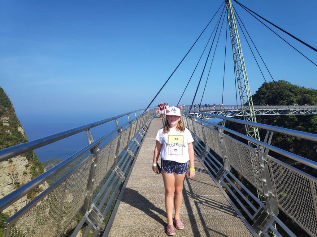 A young girl smiling at the camera while standing on a bridge in Langkawi. The sky is a beautiful blue and there is a small rocky tree-lined outcrop to the lfet and right of the picture.