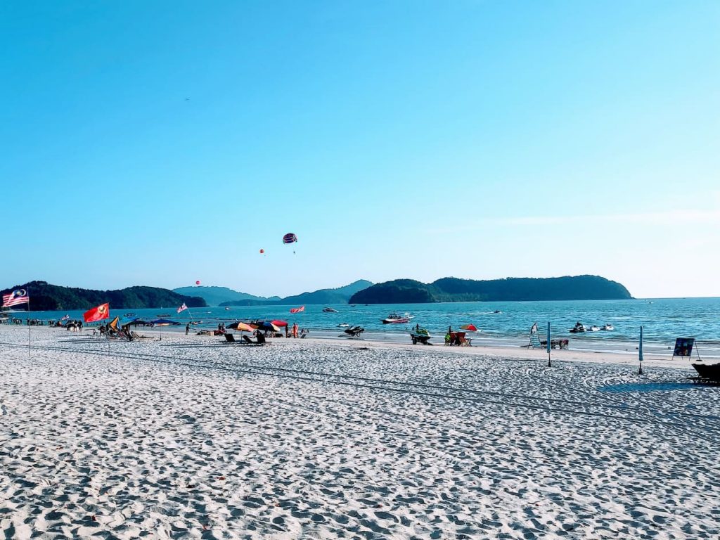 A picture of a sandy calm beach with islands in the background. On the beach, you can see some flags, chairs with umbrellas over them and some jet skis on trailers. Above the beach, there are some para-sailers in the air with colourful shutes.
