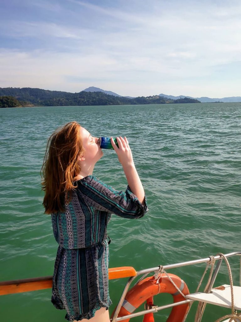 A young red-haired girl drinking from a can of soft drink on the deck of a boat. The water around her is deep green a there are tree filled island in the background