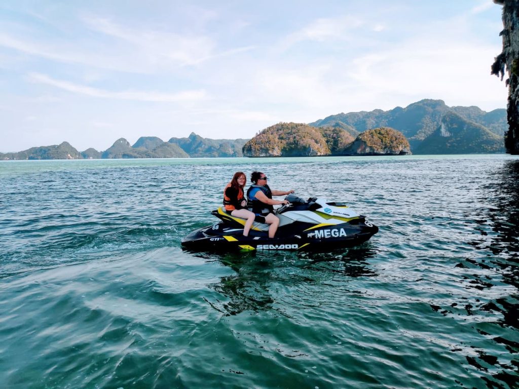 a mum and daughter on a sea doo wearing life jackets and the daughter is looking at the camera smiling while the mum concentrates. There are steep cliffs to the right with mountainous islands in the distance. The water is a deep green. Langkawi with kids is amazing.
