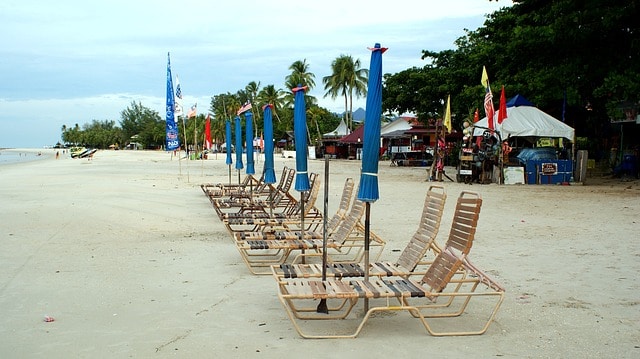 Picture of a sandy beach the sun chairs with folded up umbrellas next to them ready to be put up for the day in the forefront. In the background, you can see shops packed up under shade tents and some jet skis on the beach as well as some palm trees
