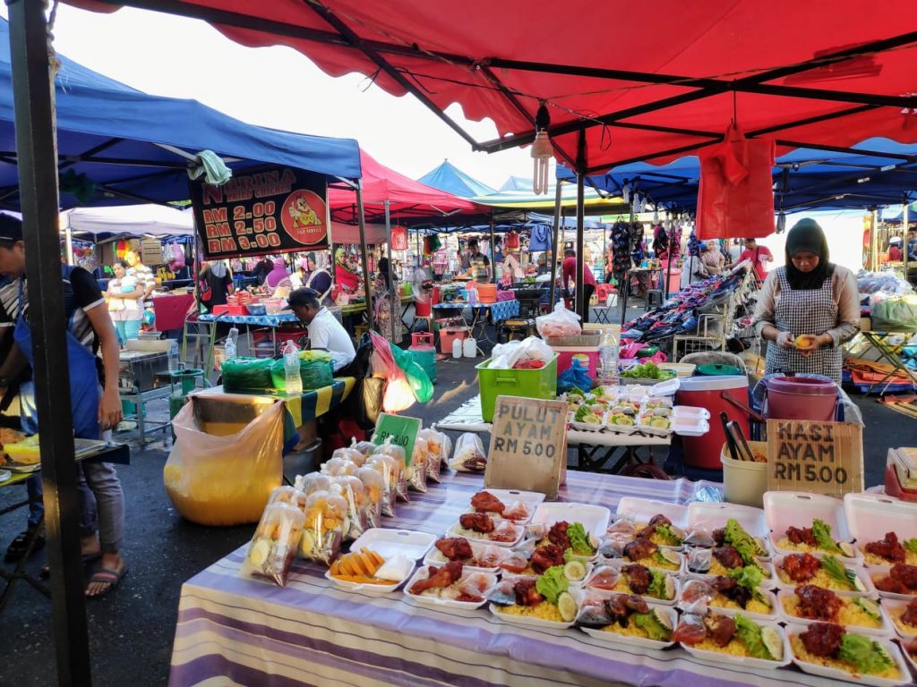 A busy market in Langkawi Malaysia. You can see stalls selling food at the front with prices put up. This one has chicken and rice with some greens on the side. The lady seems to be cutting some mango into a bin