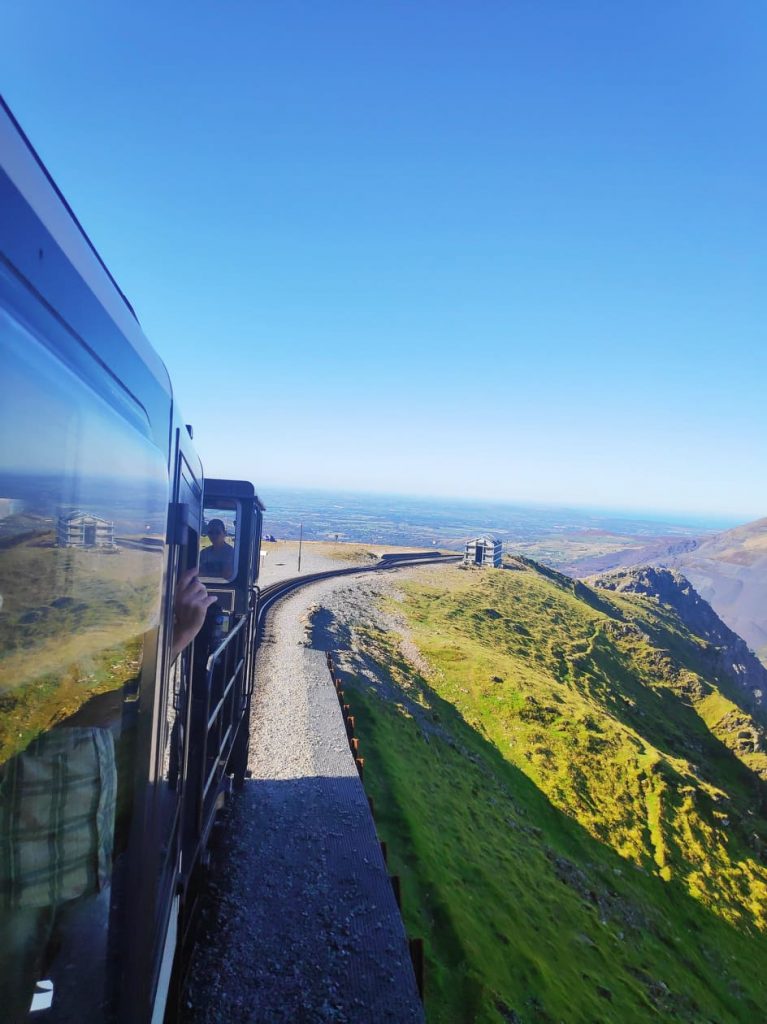 Nearing the top of Mount Snowdon on a steam train. In the background, you can see the faint trees and land below the mountain. In the foreground, you can see the lush green mountain slopes with the train tracks coming up the mountain. Riding the Snowdon Railway is one of the best things to do in North Wales