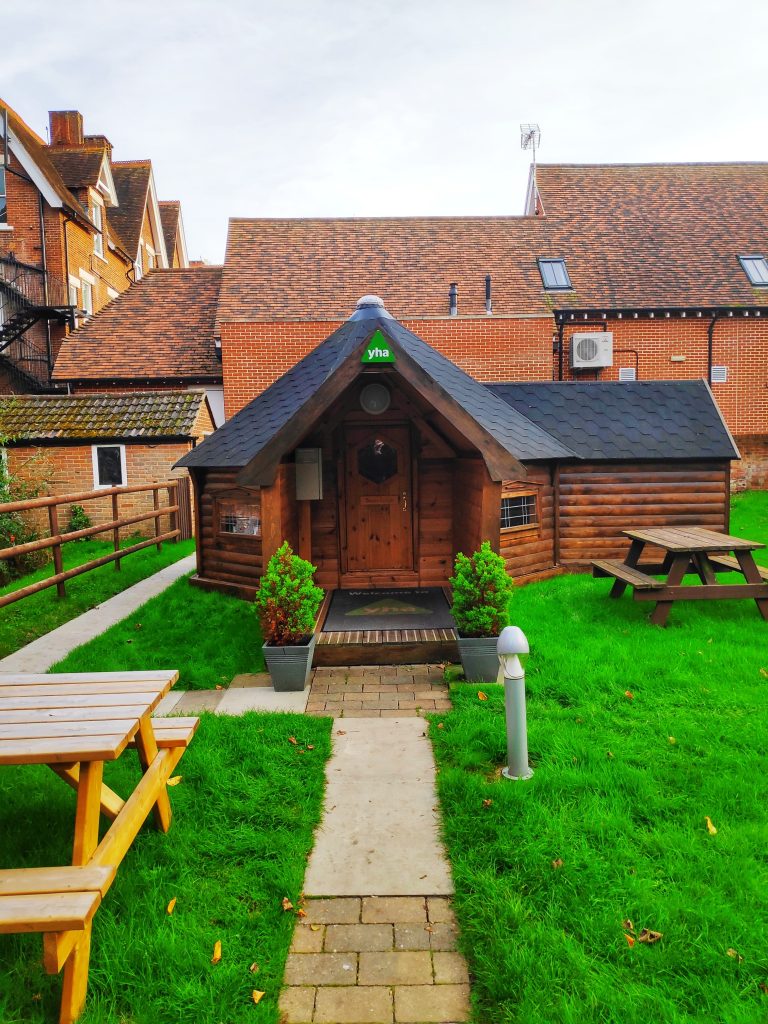 A small wooden cabin at the back of the YHA Canterbury. The path leads to the front door through the green grass. It looks like a small hobbits hut