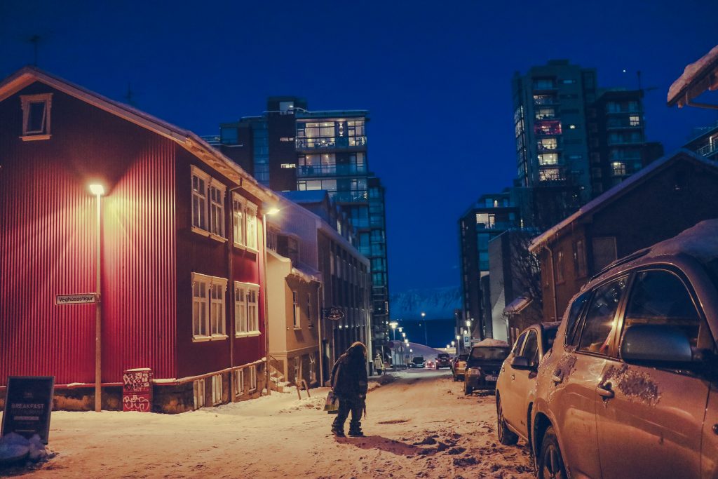 Night time in Reykjavik with snow on the ground and a person slowly crossing the road between the high rise buildings