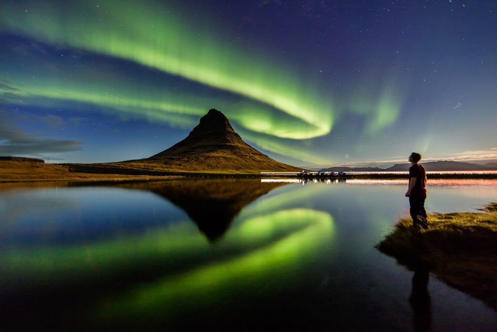 Northern lights dancing over a pyramid shaped mountain in Iceland with a man gazing up into the sky.