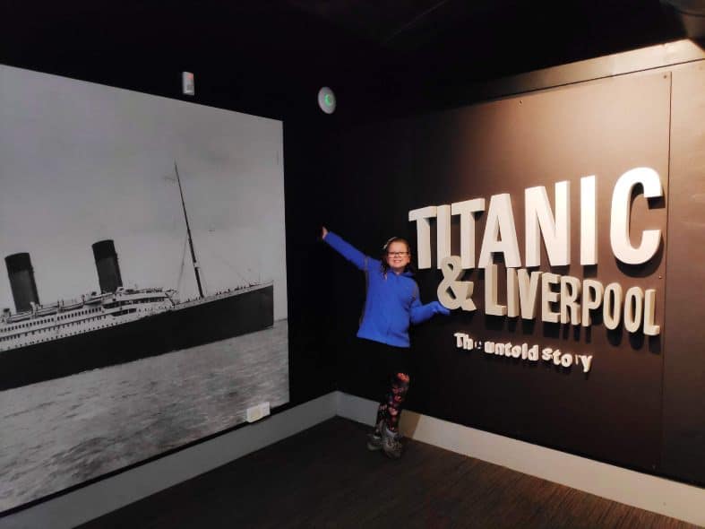 Travel with teens to Liverpool to see the Titanic story in the city. A teen looks excited to be at the museum with one arm raised and a big smile