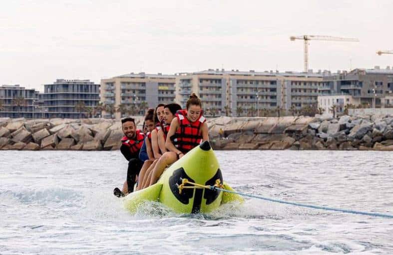 Teens and young adults on a banana shaped inflatable getting towed nehind a boat in Barcelona