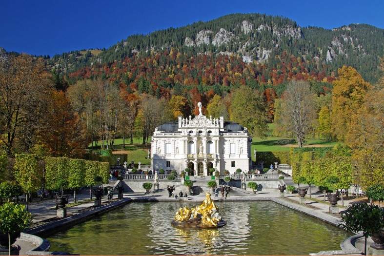 A palace in Bavaria sitting with the mountains in the background and a pond at the front