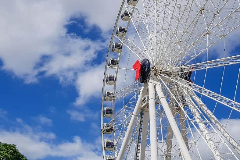 The Brisbane Ferris wheel is towering toward a cloudy sky with a blue backdrop. The pods look small compared to the white wheel. Riding the Ferris wheel is one of the best things to do in Brisbane with teenagers