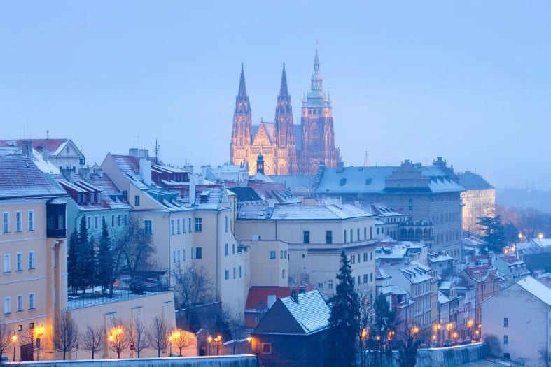 The steeples of the Prague Castle Church are lit up at night with lights. The buildings in the forefront are covered in snow with the trees bare of leaves.