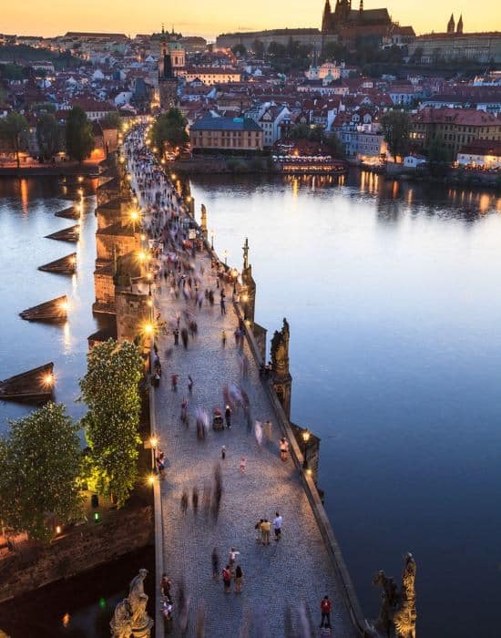 A picture of Charles Bridge in Prague from one of the towers. The bridge seems to come out of the bottom of the picture and stretches to the top of the frame The picture is taken at dusk with the lights on the bridge and the nearby buildings reflecting off the bridge and the water. You can see Prague castle high on the hill in the distance