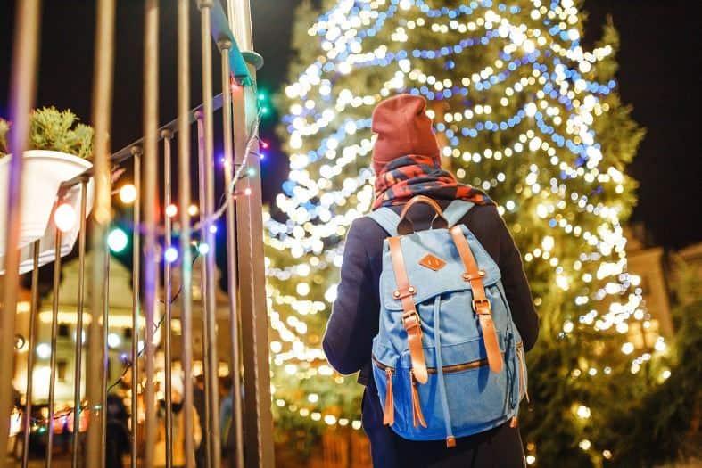 A teen with a red beanie on and a blue backpack is wandering a Christmas market. There is a big decorated Christmas treet with silver and blue lights on it in front of them