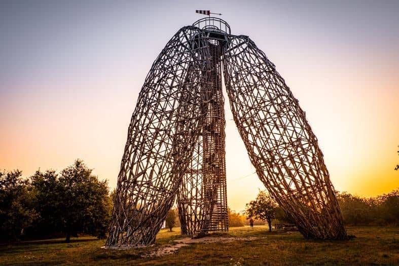 This sculpture is of 3 egg-shaped columns that meet at the top at their points. They look like they are made from woven reeds with an enclosed spiral staircase in the middle that takes you to the top. The sunset in the background is beautiful lighting up with sky and the surrounding trees with a golden colour.