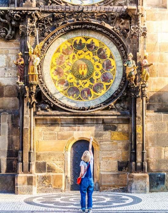 A teen standing pointing to the Prague Astronomical Clock in a set of blue denim overalls. The tiles in front of them are black and white in colour. The Astronomical clock is yellow in colour and has 11 smaller paintings surrounding a central one in the middle of the clock's face. There are also sculptures on the side of the clock and a small black door under it making it one of the things to do in Prague