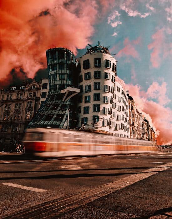 The dancing house of Prague is a building that seems to lean on the one beside it. It is a dark grey colour with large black windows and it looks like a cage it surrounding it. The one next to it is white with large black windows. There is a blurred train passing by the front of the building and the picture has been edited to make the clouds in the sky above it a redish pink colour.