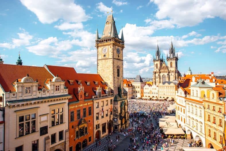 A picture of the Prague Old Town taken from one of the towers nearby. There are plenty of people wandering the old town of Prague. The sun is shining bright on the buildings highlighting the reds, oranges and creams that they are painted in. you can see the church spires in the distance