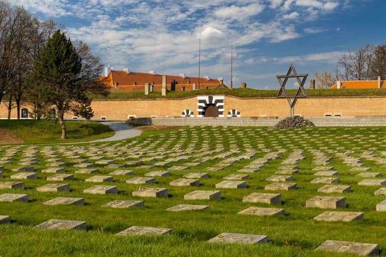 In the forefront of the picture are small concrete square gravestones with a Star of David to the right of them. Behind that is a brick wall that runs the length of the picture with buildings at Terezin Camp in Czech Republic appearing over the top of the wall. Things to do in Prague with teenagers.