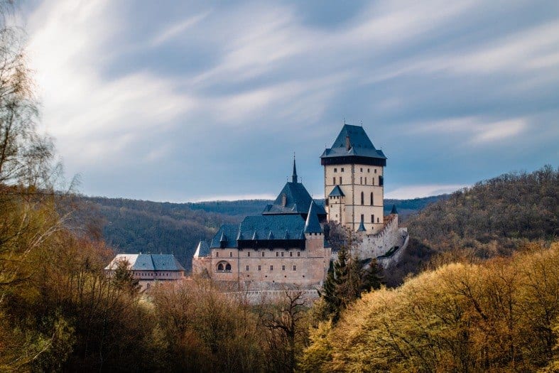 Karlstejn Castle is in the countryside just out of Prague. The trees around the castle are yellow in colour and starting to lose their leaves. The castle is an off white colour with its largest tower standing taller than the others. The rood is a royal navy blue