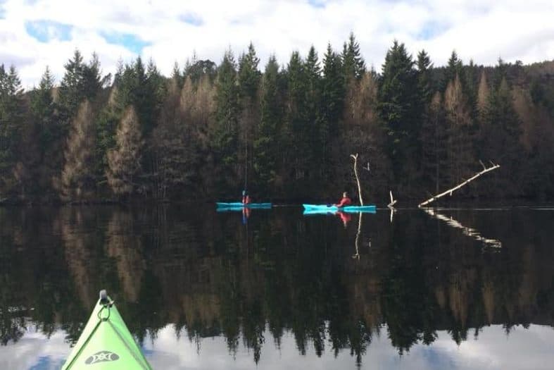 Canoeing on the lake in Scotland is a great teens adventure activity. 2 light blue canoes glide in a still lake the reflection of the green trees coming off the water. Some trees are still dark green while others have lost all of their leaves and seem to just be bare and just branches.