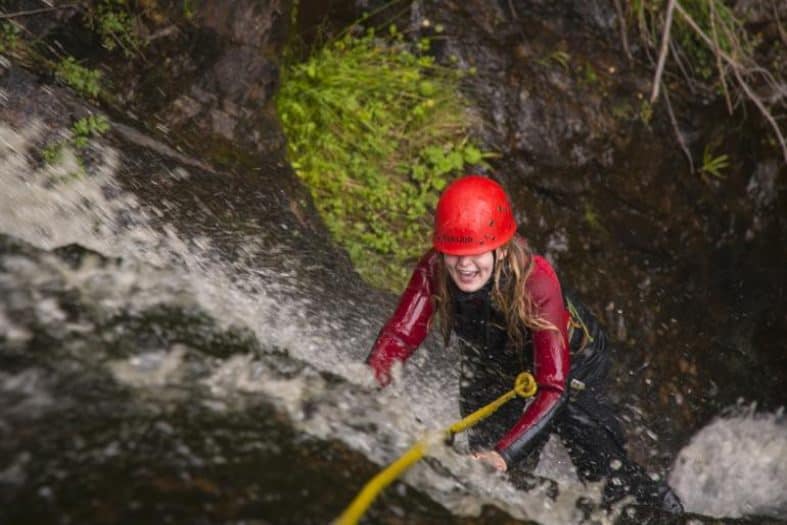 A young girl in a red wetsuit climbs up a fast-flowing waterfall with a big smile on her face. A yellow rope is guiding her and her red hard hat is covering her eyes as she gets completely drenched from the water on her teens adventure holiday