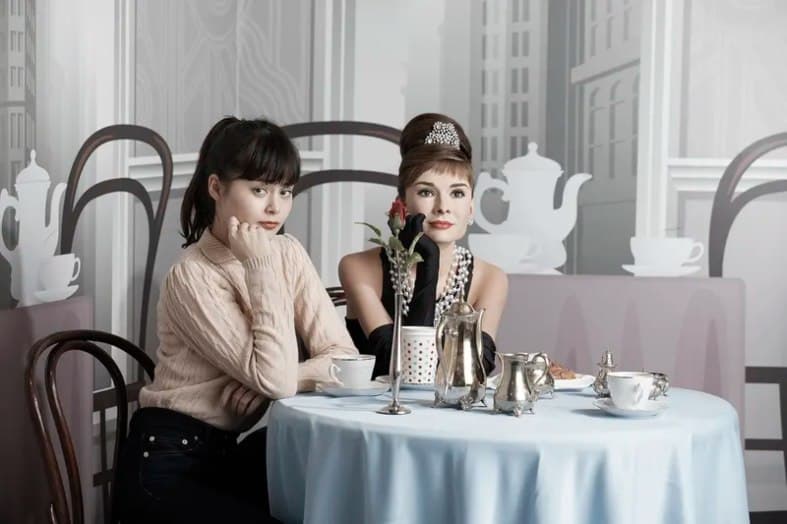 a girl sitting with a wax model of audrey hepburn at madame tussauds