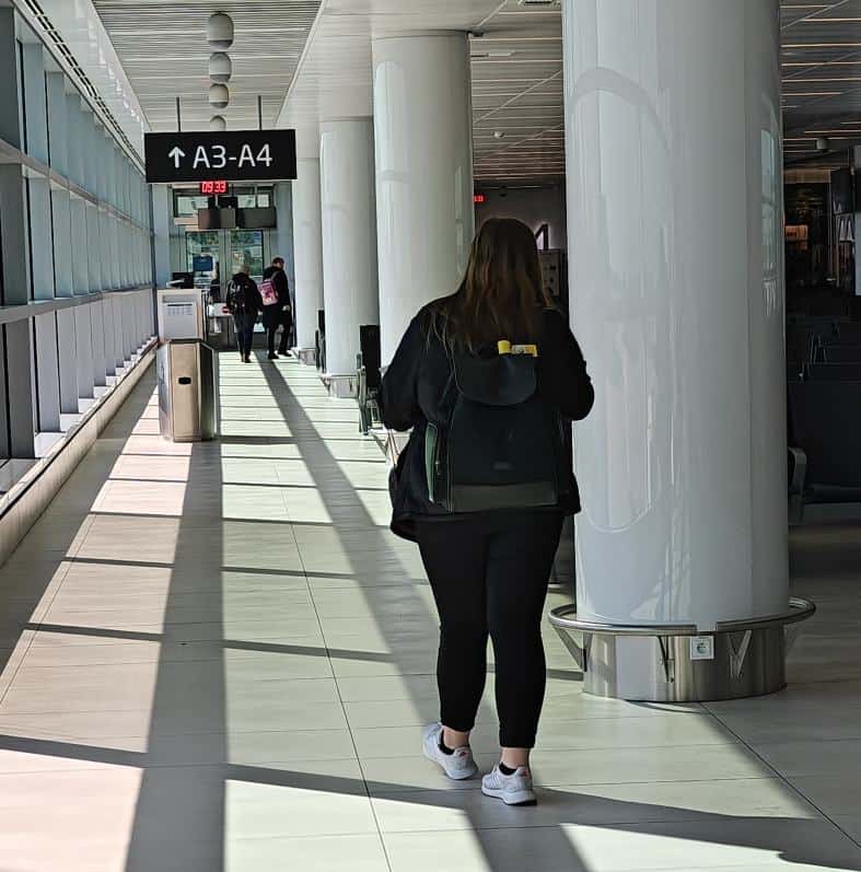 A teenager walking through the Frankfurt Airport with a backpack on her back. She is dressed in comfortable clothes and is walking next to a long window with plenty of sunlight on the floor towards gate A3-A4. long haul travel with teenagers
