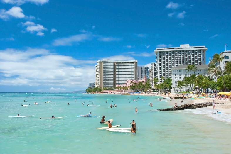 Tourist enjoying the beach at Waikiki. You can see some clouds in the sky and some apartment buildings in the background. The water is calm and a clear blueish-green colour. Vising the beach is one of the best things to do in Honolulu with teenagers