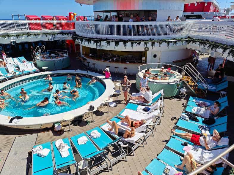 Pool area on Virgin Voyages' ship, featuring lounge chairs, umbrellas, and passengers relaxing by the water on a sunny day.